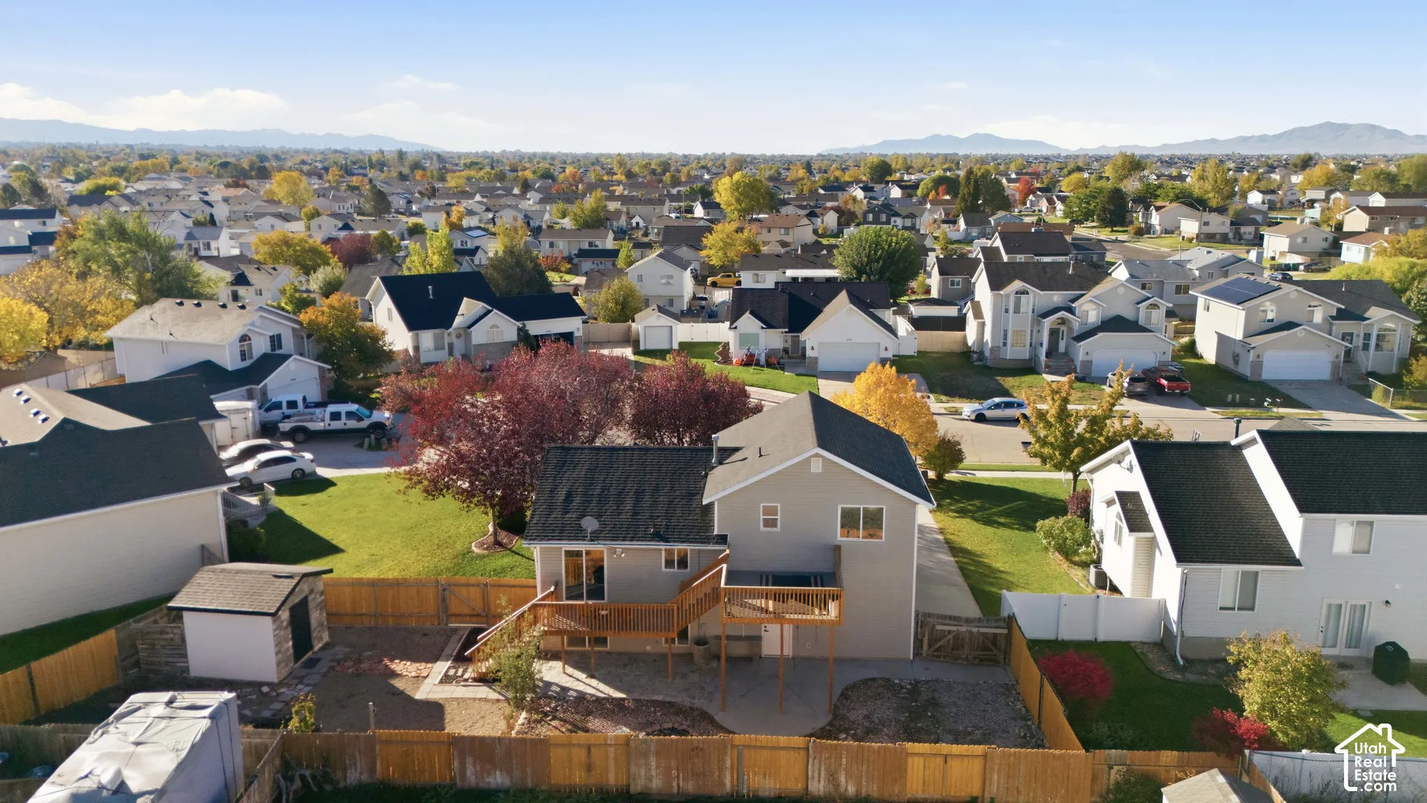 Aerial perspective of a multi-level residence featuring a wooden deck, stone-accented detached storage structure, fully fenced perimeter, and mountainous horizon