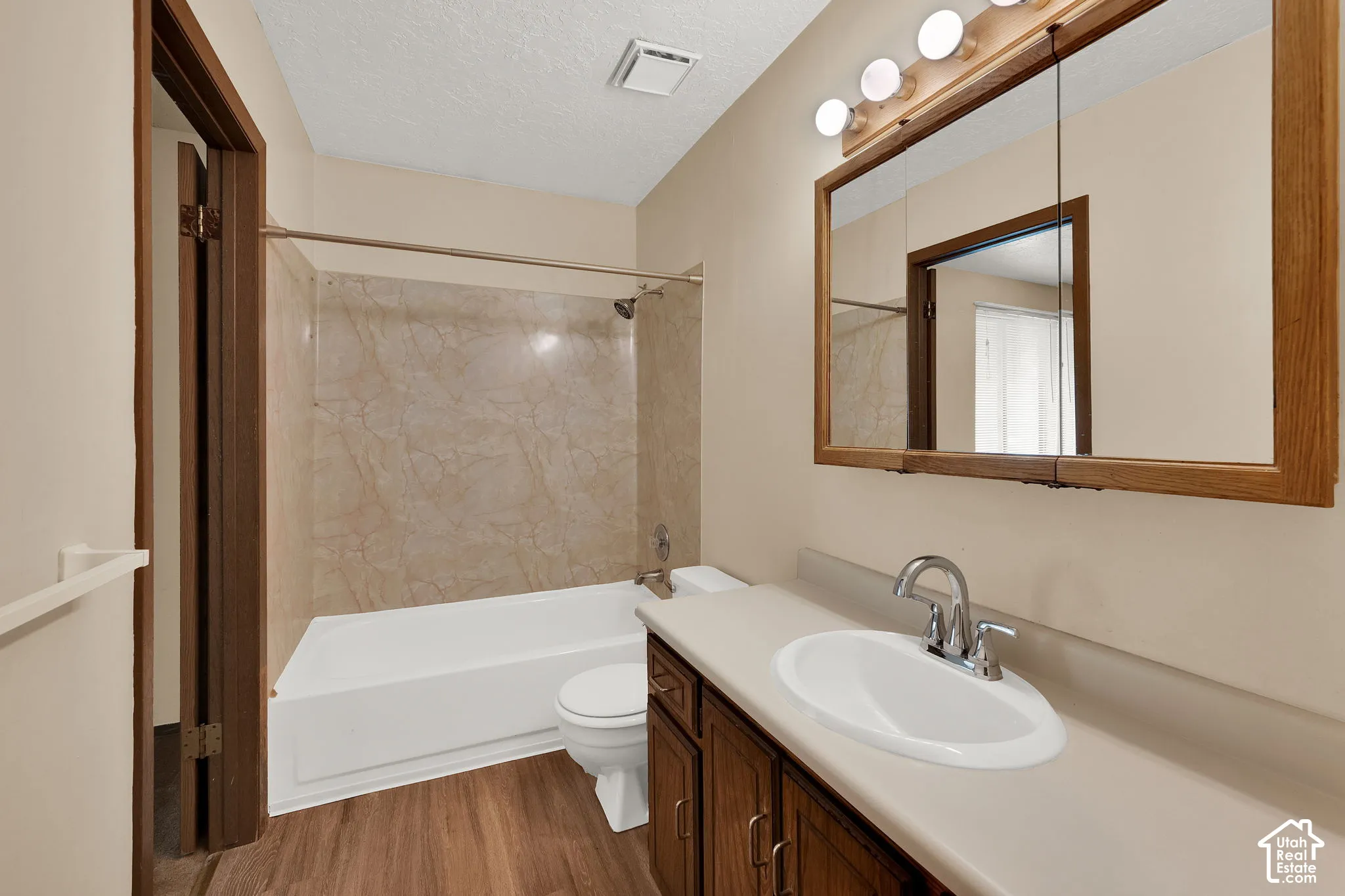 Bathroom featuring wood-finish flooring, vanity with integrated basin, framed wall-mounted mirror, four-light fixture, and shower-tub combination with surround paneling