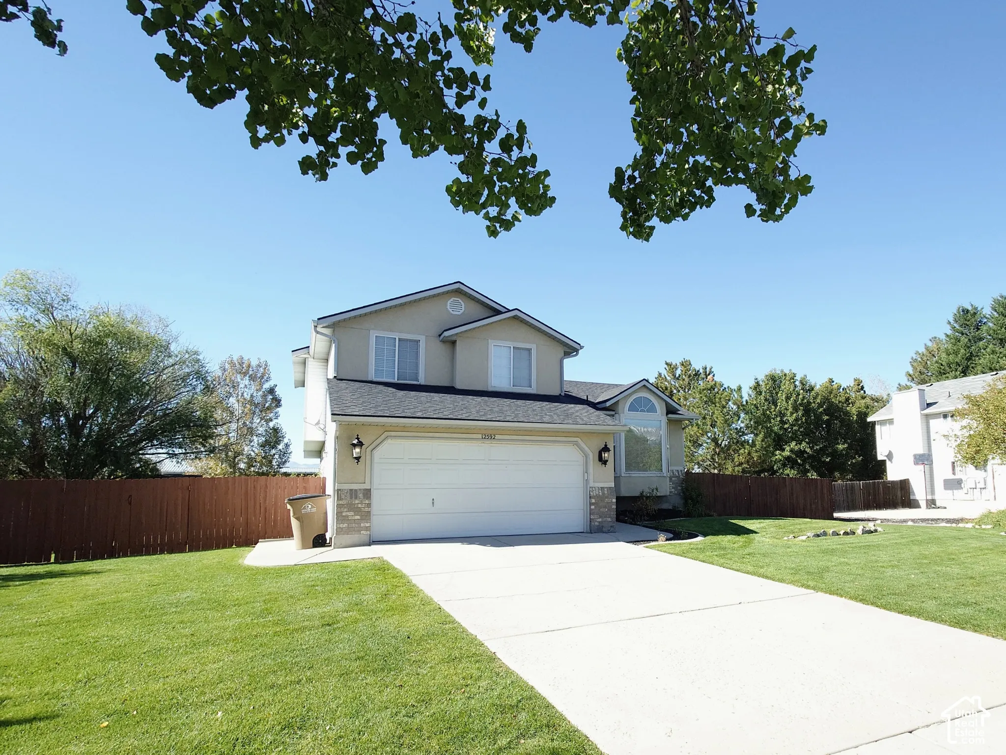 Two-story exterior featuring tan siding, a stone veneer base, a dual-car garage, and a concrete driveway surrounded by a manicured lawn and perimeter fencing