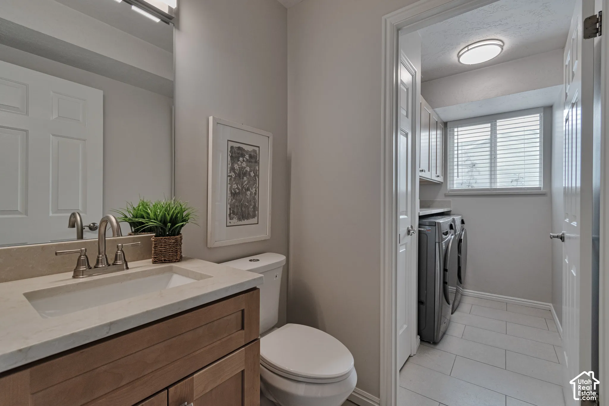Vanity featuring a light wood finish, neutral countertop, and brushed metal faucet, complemented by a laundry area with tile flooring, built-in upper cabinetry, and a window with blinds