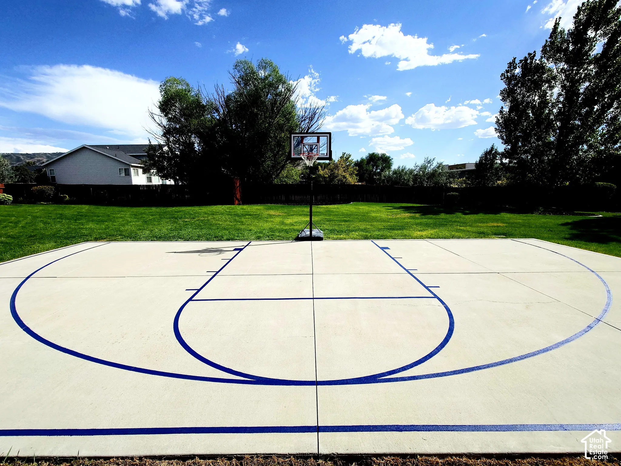 Concrete sports court with painted game lines and a backboard system, situated within a fenced backyard featuring a grass lawn and perimeter trees