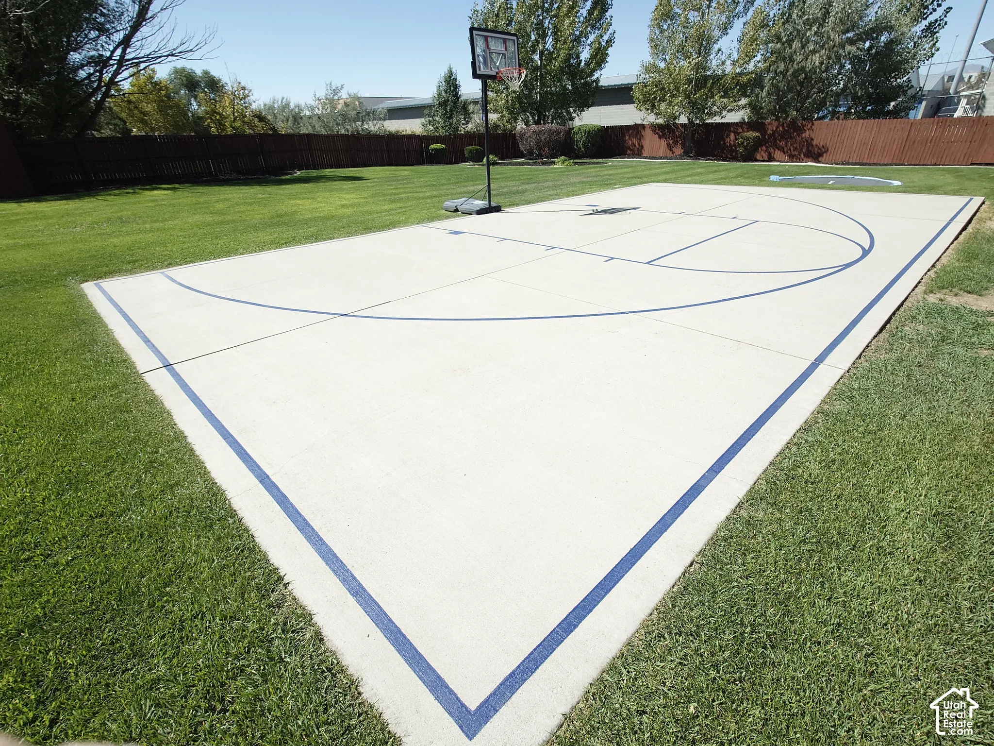 Concrete basketball court featuring painted lines, surrounded by a manicured lawn and perimeter wood fencing