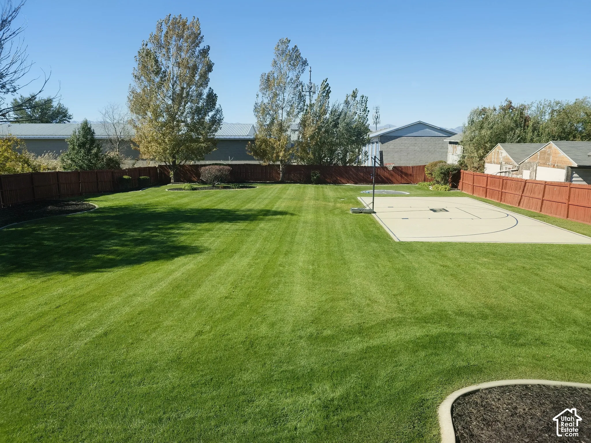 Expansive backyard featuring a large grass lawn, a paved sports court, mature trees, and perimeter wood fencing