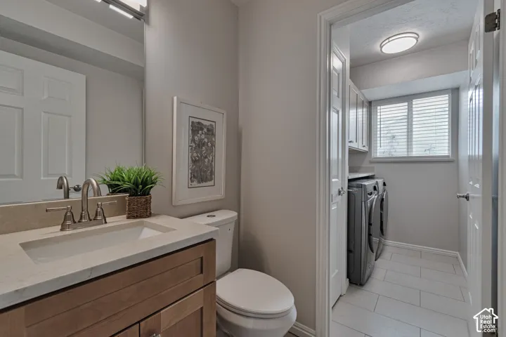 Vanity featuring a light wood finish, neutral countertop, and brushed metal faucet, complemented by a laundry area with tile flooring, built-in upper cabinetry, and a window with blinds