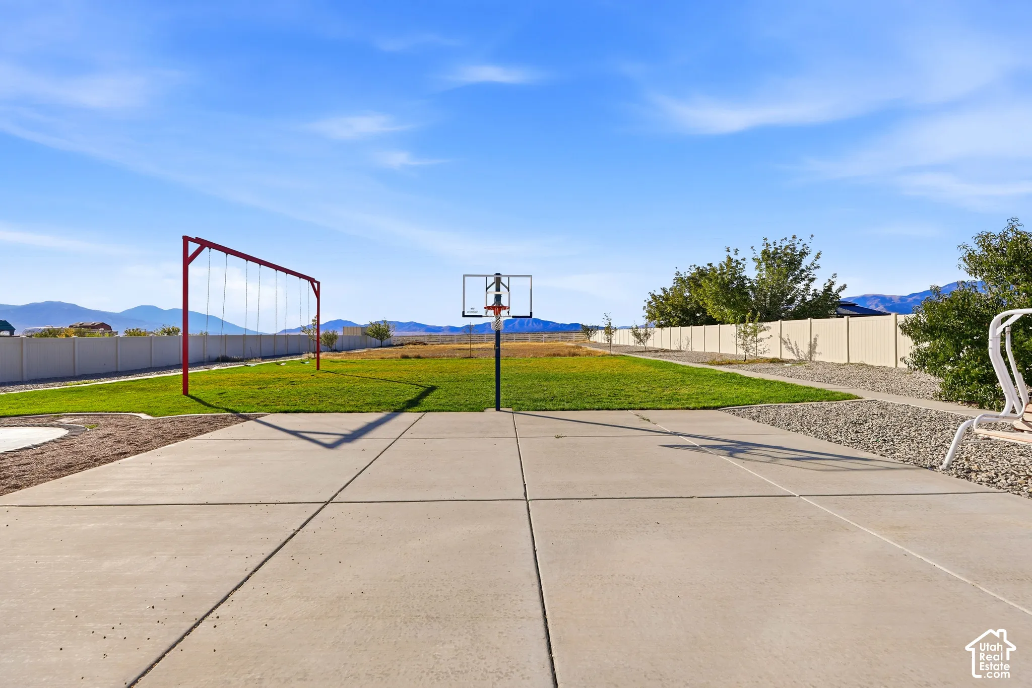 Expansive backyard featuring a concrete sport court, basketball hoop, swing set, manicured lawn, and full-perimeter vinyl fencing with mountain backdrop
