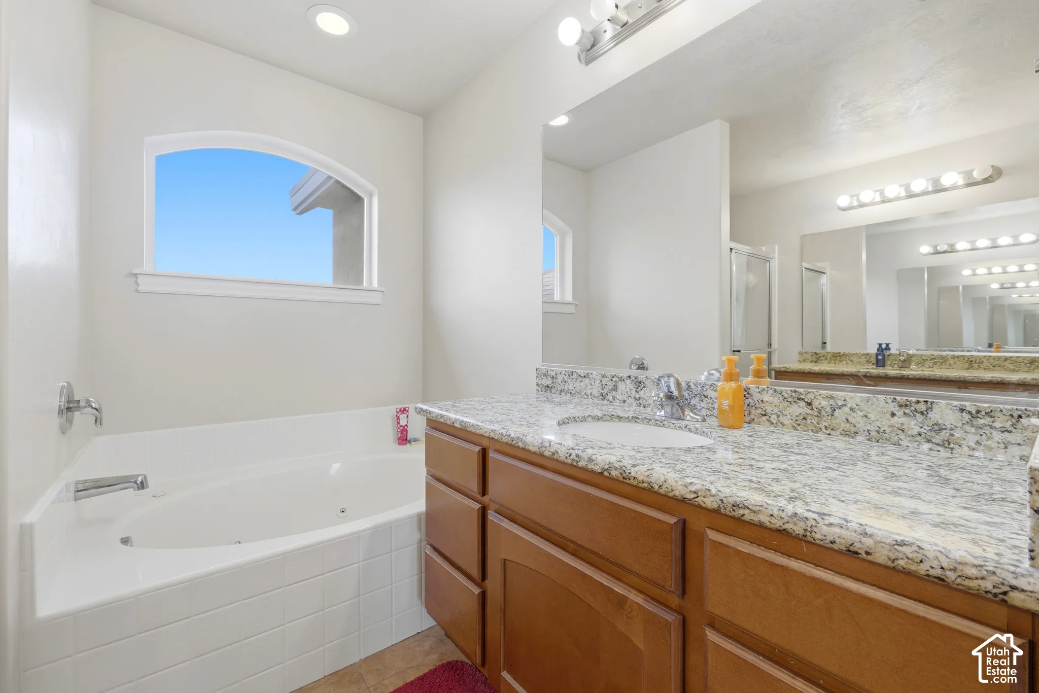 Jetted soaking tub with tile surround, vanity with stone-finish countertop, undermount sink, and large wall-mounted mirror