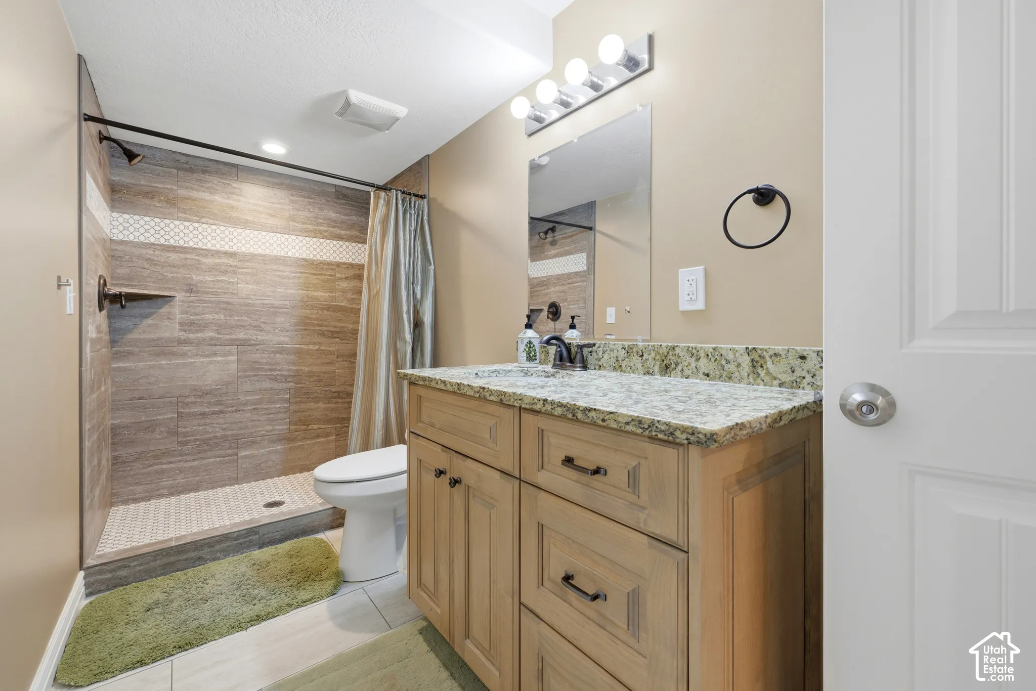 Bathroom featuring a wood-tone vanity with stone countertop, drop-in sink, walk-in shower with tiled surround, and multi-bulb wall lighting