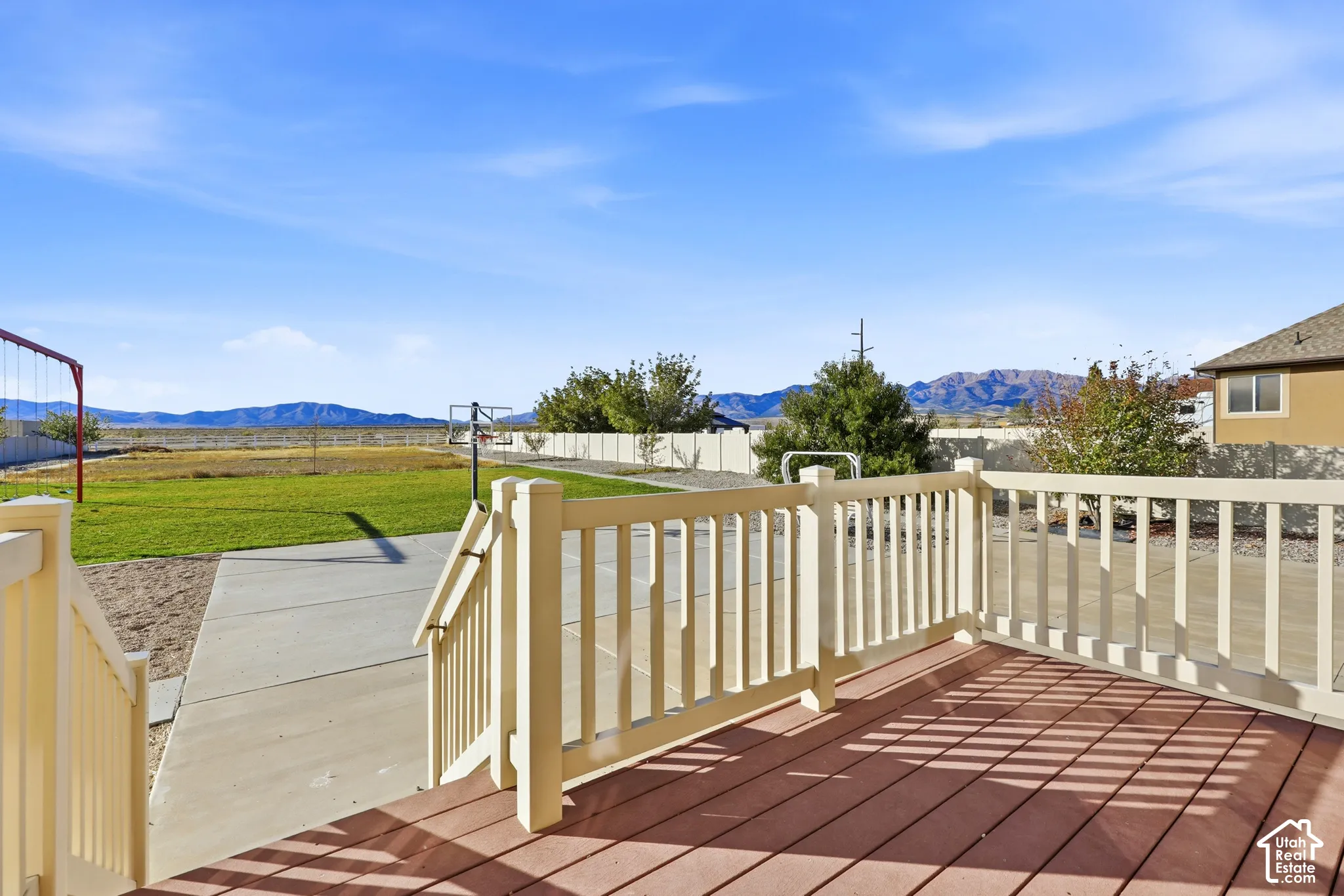 Composite wood-finish deck with cream-colored railing, expansive concrete patio, manicured lawn, perimeter fencing, and mountain backdrop