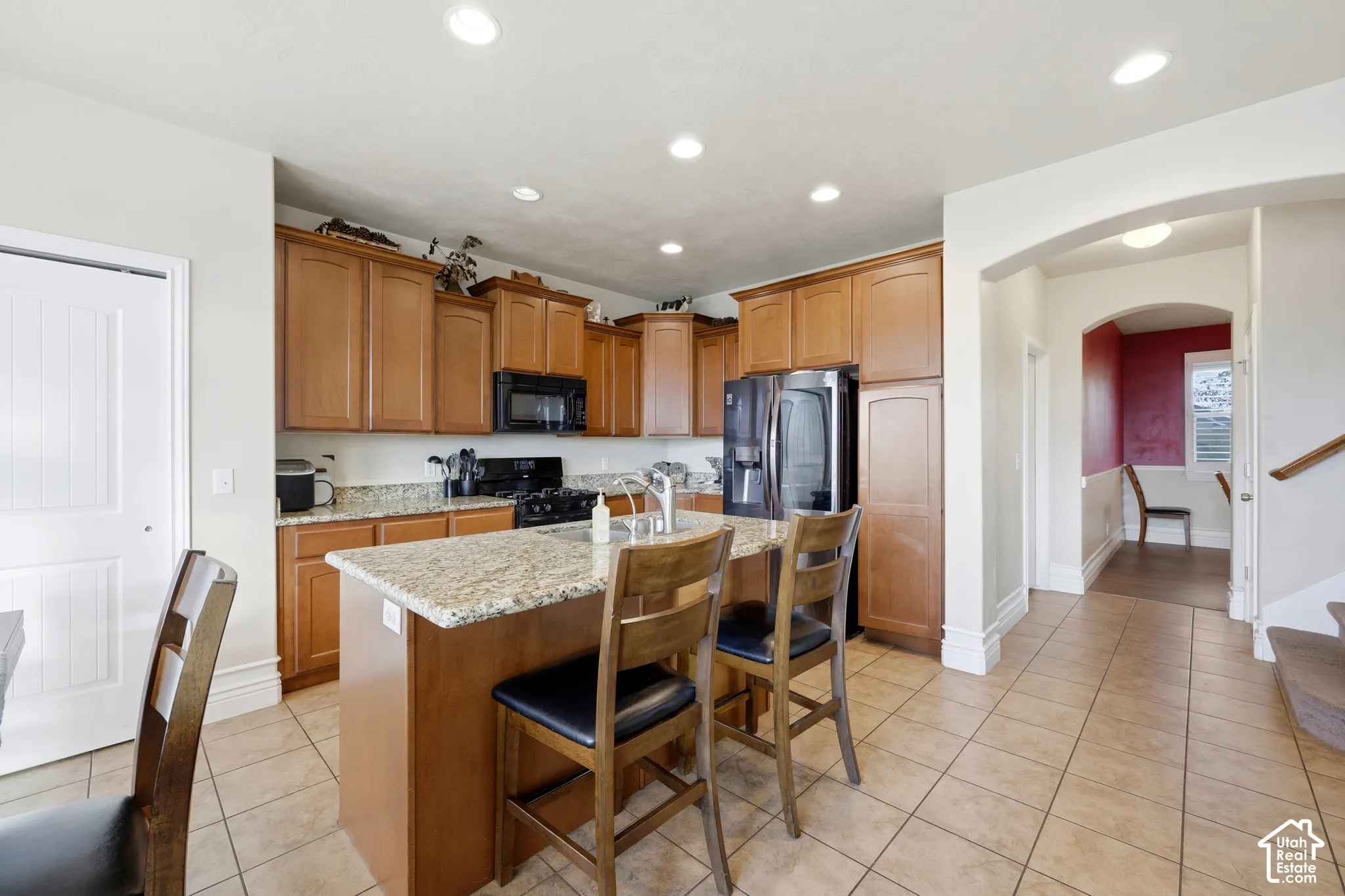 Kitchen featuring medium-toned wood cabinetry, stone-surface countertops, a center island with an integrated stainless steel sink, recessed lighting, and ceramic tile flooring