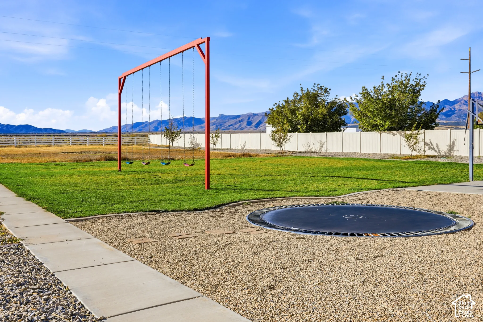 Expansive backyard featuring a grass lawn, gravel grounds, a metal swing set, an in-ground trampoline, and mountain scenery