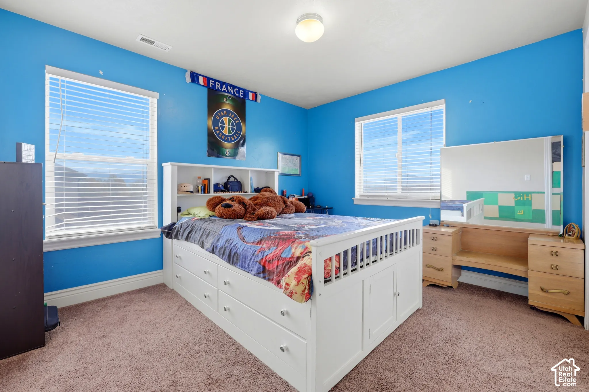 Dual-aspect room featuring neutral carpeting, two windows with horizontal blinds, a white built-in bed frame with integrated storage drawers, and a built-in wooden vanity with a large mirror