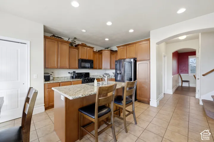 Kitchen featuring medium-toned wood cabinetry, stone-surface countertops, a center island with an integrated stainless steel sink, recessed lighting, and ceramic tile flooring