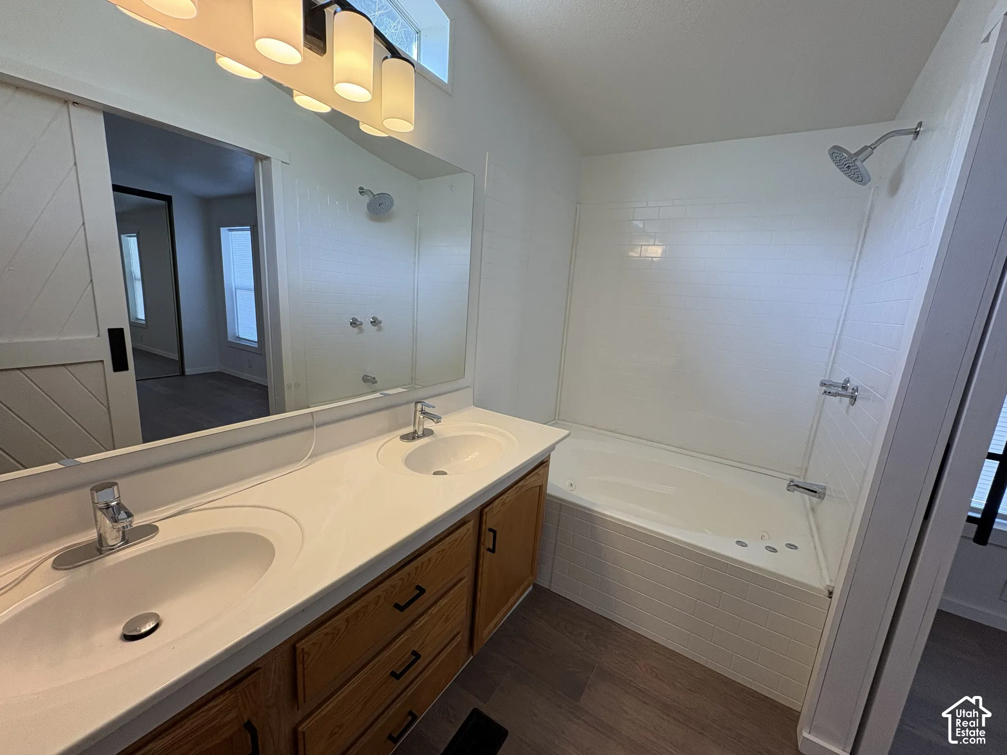 Dual-sink vanity with a solid-surface countertop, white subway tile tub surround, chrome fixtures, and wood-finish flooring
