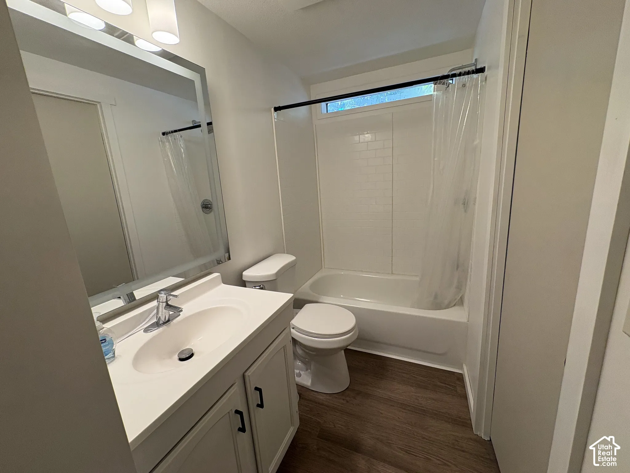 Bathroom featuring wood-finish flooring, a white vanity with an integrated sink, a large rectangular mirror with integrated lighting, and a bathtub with a white surround and transom window