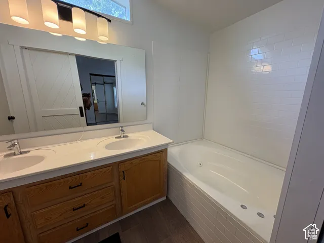 Dual-sink vanity with light wood-finish cabinetry, solid surface countertop, jetted soaking tub, white subway tile surround, and horizontal five-light fixture