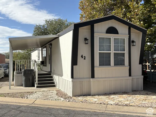 Exterior featuring light-toned vertical siding with contrasting dark trim, attached metal carport, staircase with railing, and decorative stone-finish ground cover