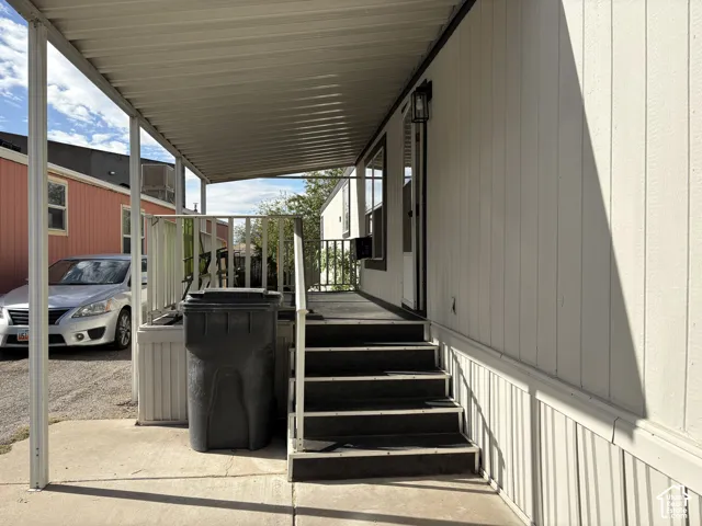 Exterior entry featuring a covered carport, raised porch with metal railing, concrete landing, and exterior wall-mounted light fixture