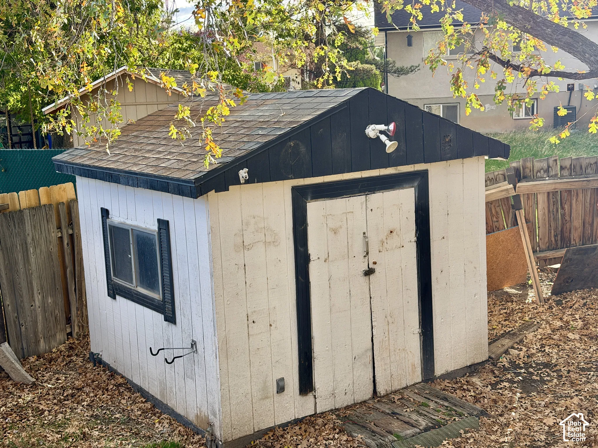 Storage structure featuring dual swinging doors, a shingled roof, a horizontal sliding window, and an exterior mounted light fixture