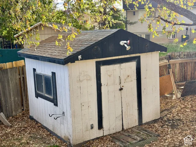 Storage structure featuring dual swinging doors, a shingled roof, a horizontal sliding window, and an exterior mounted light fixture