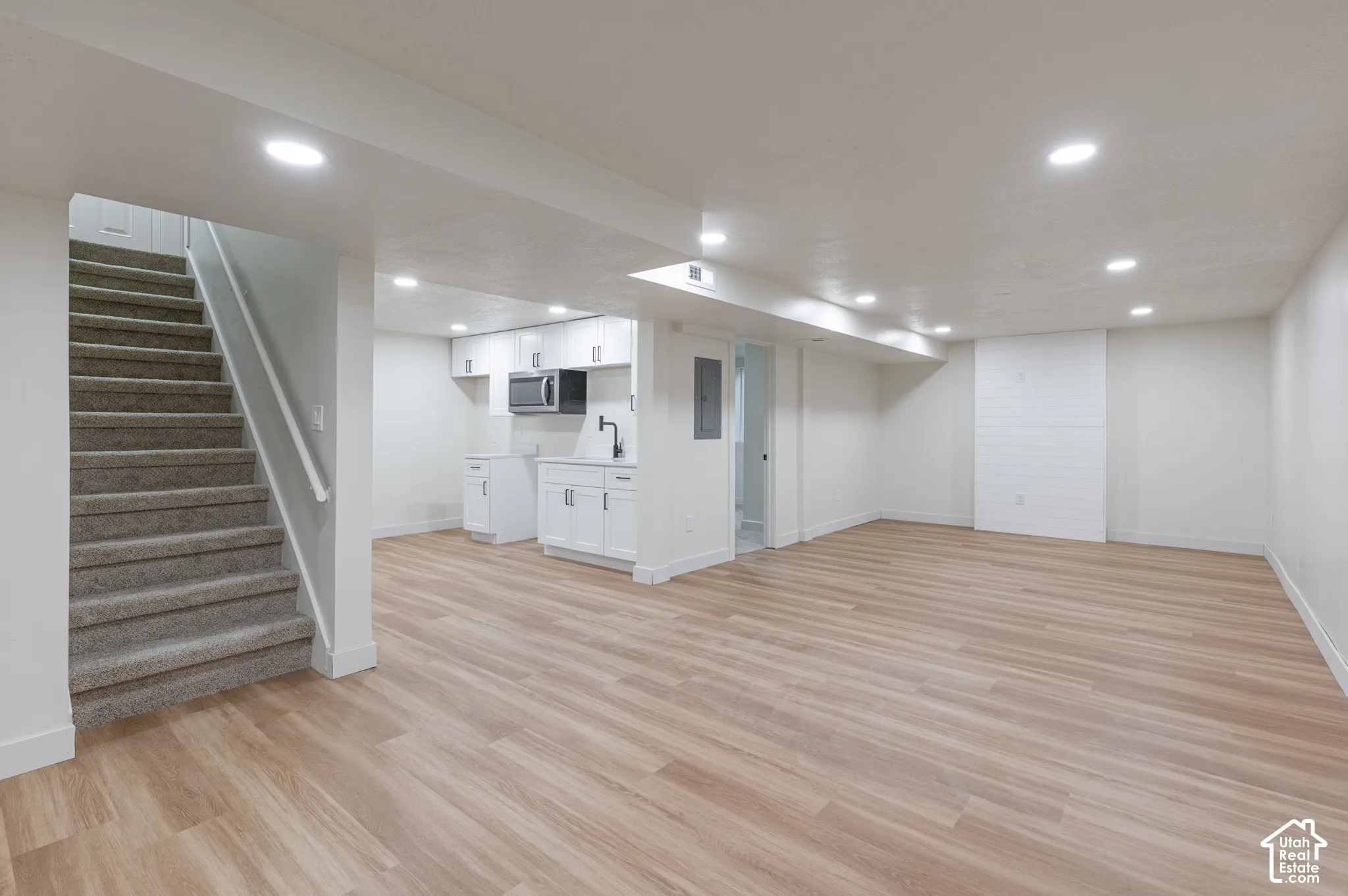 Open-concept interior featuring light wood-finish flooring, recessed lighting, carpeted stairs, and a kitchenette with white cabinetry and a built-in microwave