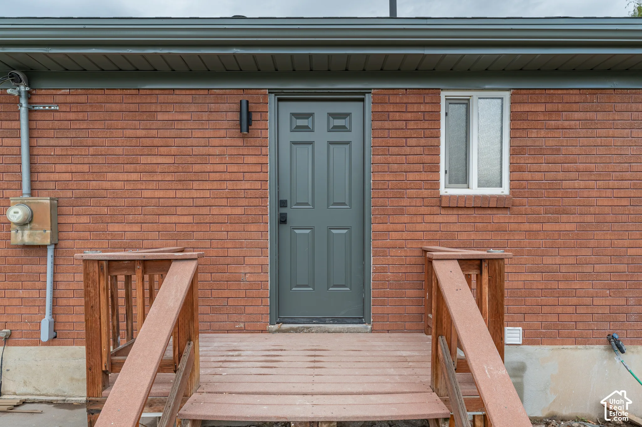 Exterior featuring red brick masonry, a wood-decked porch with railing, a dark-toned panel door, and a wall-mounted light fixture