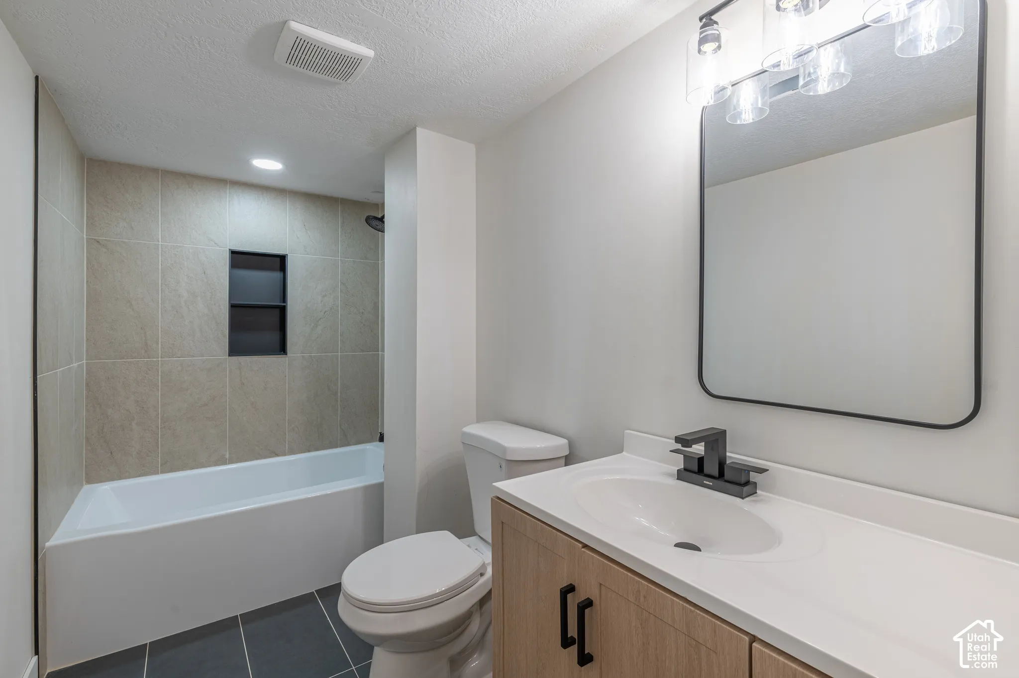 Bathroom featuring a wood-finish vanity with matte black hardware, integrated white countertop sink, rectangular wall-mounted mirror, and a tile-surround bathtub with a built-in recessed niche