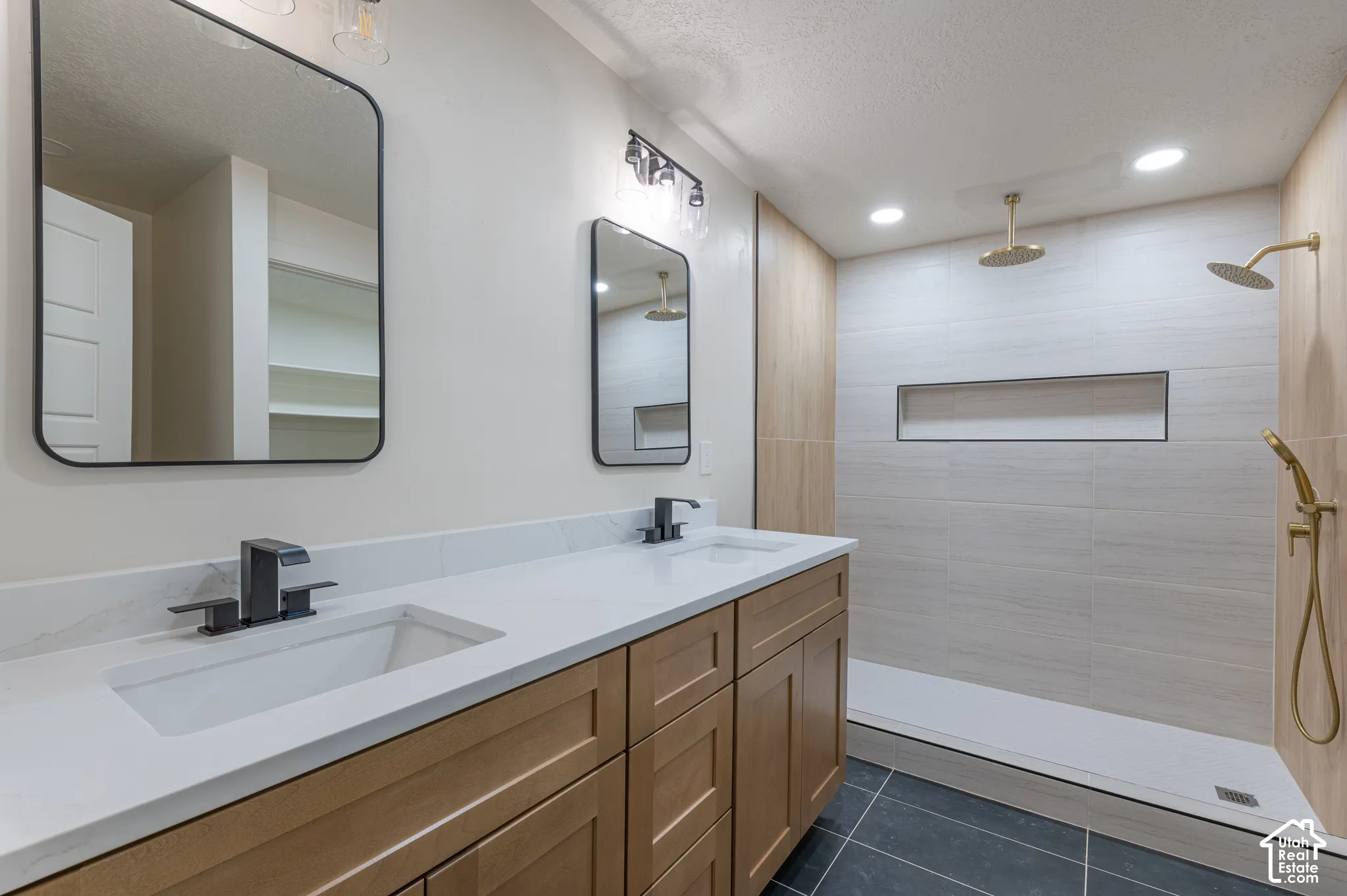 Dual-vanity cabinetry with a quartz countertop, rectangular undermount sinks, matte black faucets, and large-format tiled shower enclosure featuring gold-tone fixtures and an inset wall niche