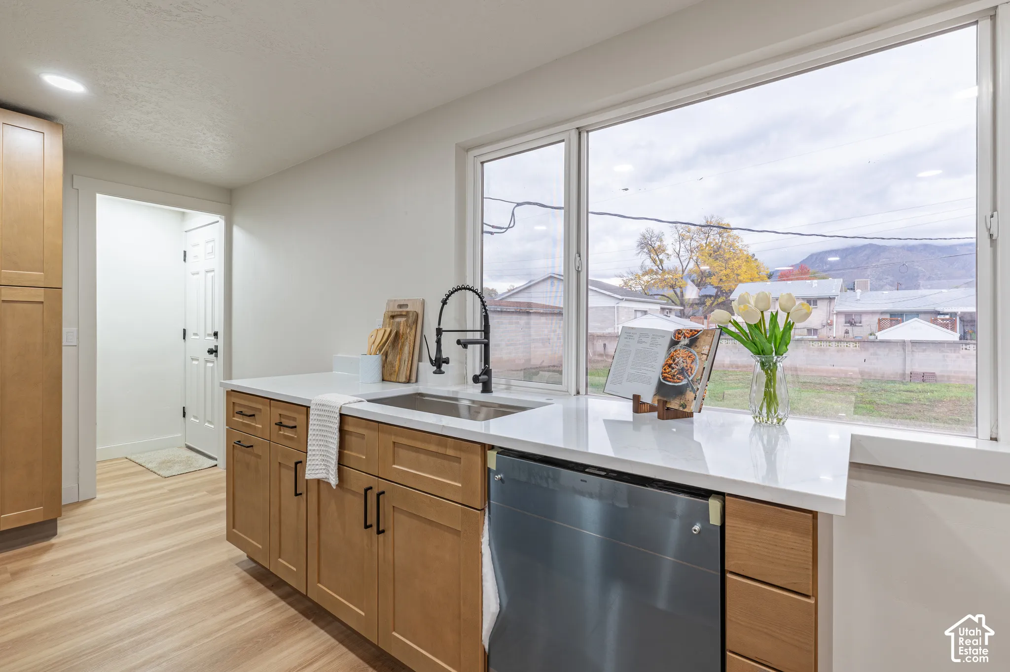 Kitchen featuring light wood-finish cabinetry, solid surface countertops, a stainless steel dishwasher, an undermount sink with a matte black high-arc faucet, and wood-finish flooring