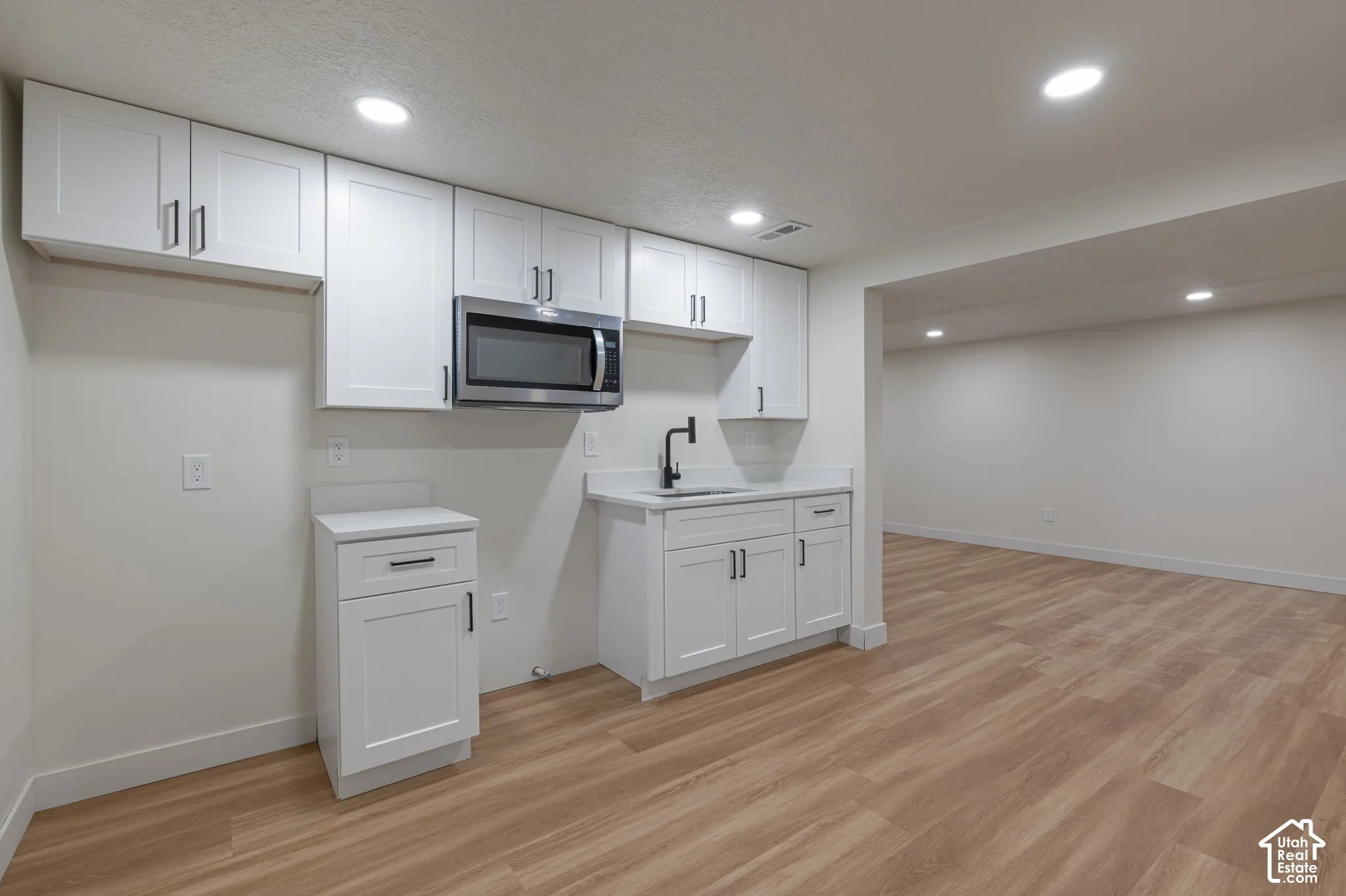 Kitchenette featuring shaker-style cabinetry, wood-finish flooring, a stainless steel over-the-range microwave, a solid-surface countertop with an integrated sink, and a matte black faucet