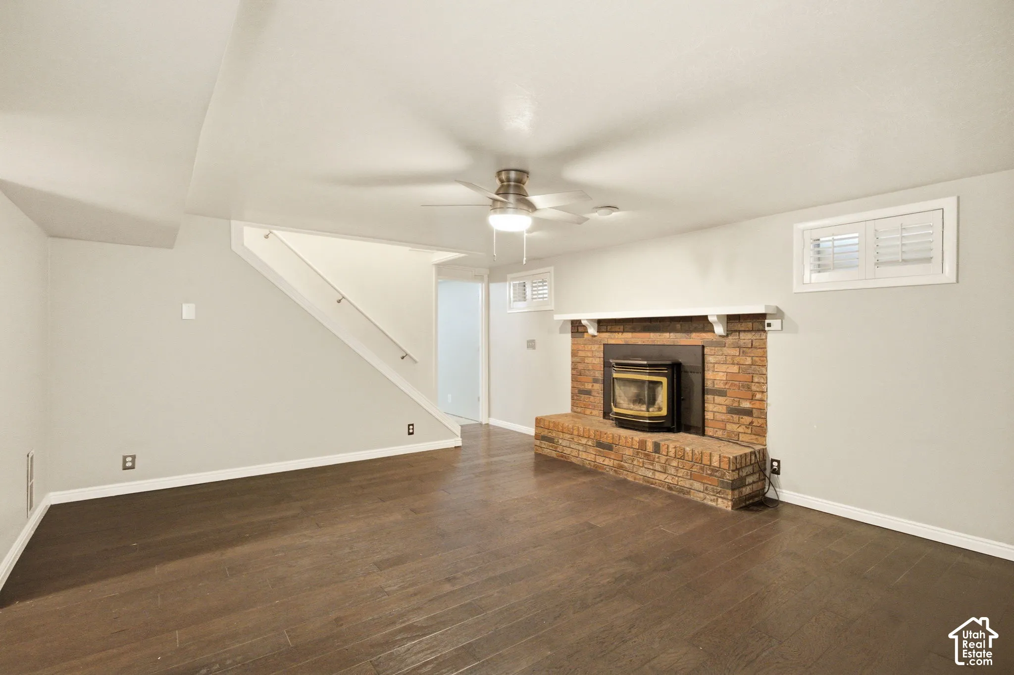Wood-finish flooring, brick fireplace with a hearth and mantel, integrated wood-burning insert, ceiling fan with integrated lighting, and white window shutters