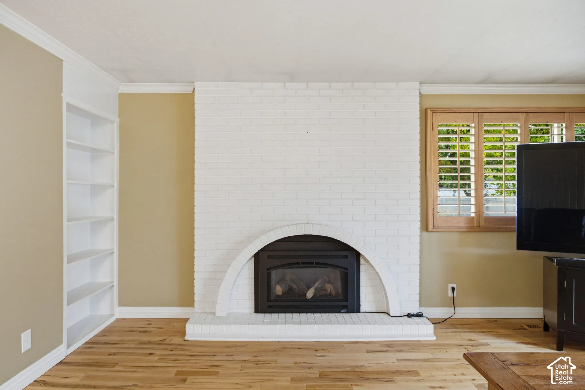 White painted brick fireplace featuring an arched surround, wood-finish flooring, built-in recessed shelving, and louvered wood shutters