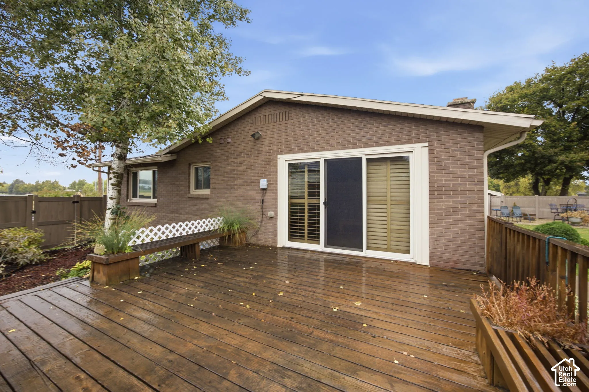 Expansive wood-finish deck featuring built-in bench seating, brick exterior, sliding glass door, and wooden perimeter fencing