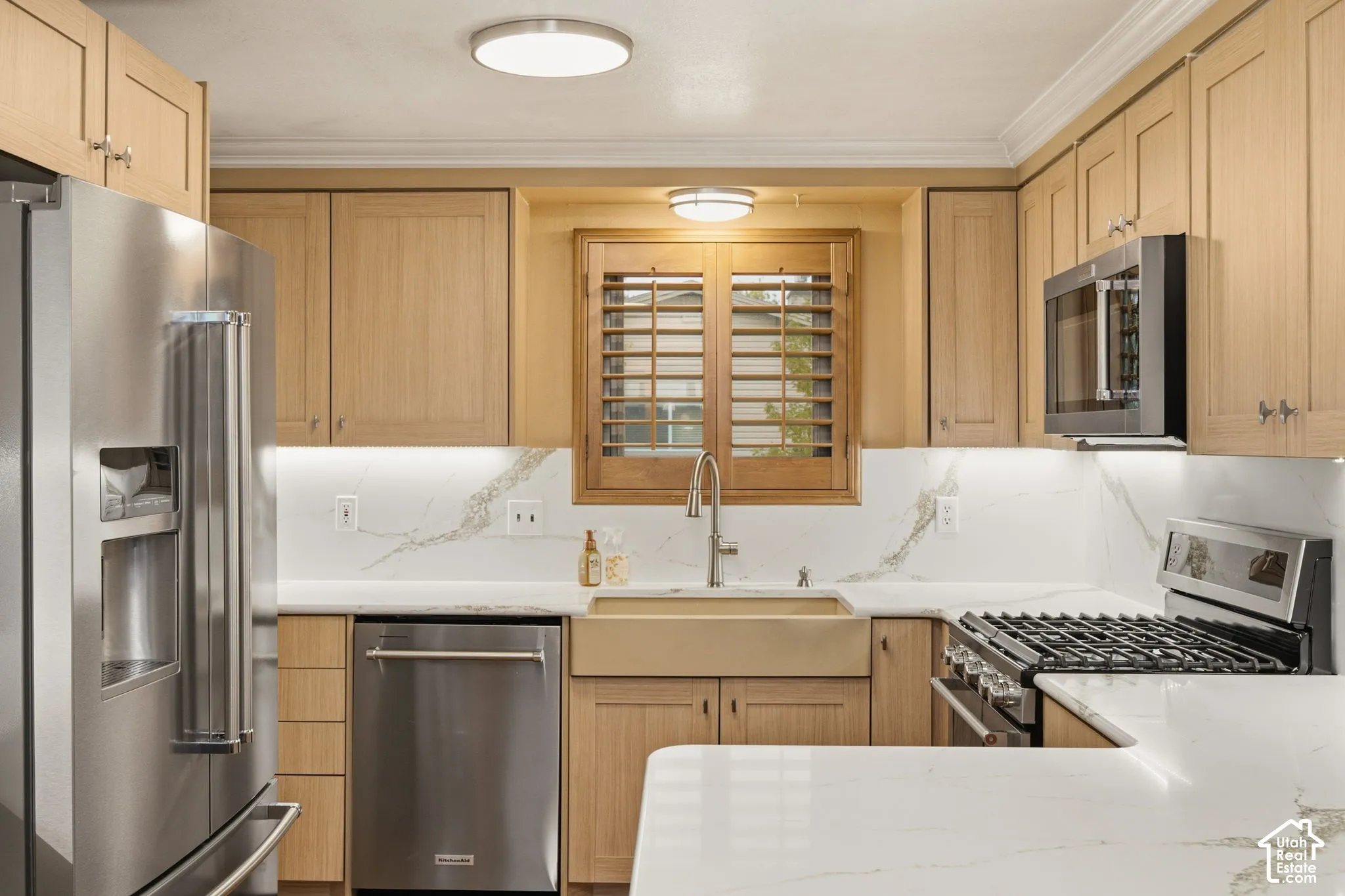 Kitchen featuring light wood-finish cabinetry, stone-patterned slab backsplash, apron-front sink with a gooseneck faucet, stainless steel appliances, and integrated under-cabinet lighting