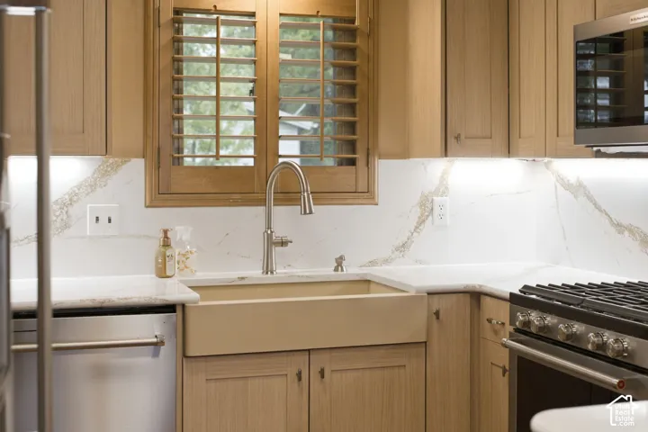 Custom wood cabinetry, apron-front farmhouse sink, stone-finish slab backsplash, stainless steel appliances, and integrated under-cabinet lighting