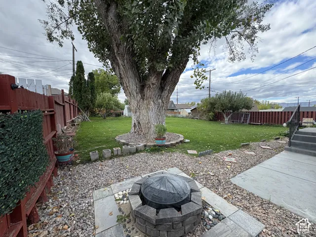 Backyard featuring a stone-encased fire pit, expansive lawn, mature tree, stone-tile patio area, and privacy fencing
