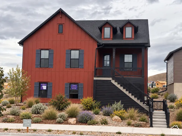 Two-story exterior featuring vertical board-and-batten siding, dark-toned shutters, a covered porch with iron railings, gabled dormers, and xeriscaped front grounds