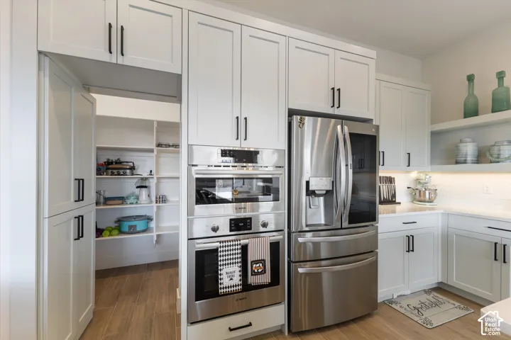 Kitchen featuring white shaker-style cabinetry, stainless steel wall-mounted oven and microwave, stainless steel refrigerator, integrated pantry with shelving, and wood-finish flooring