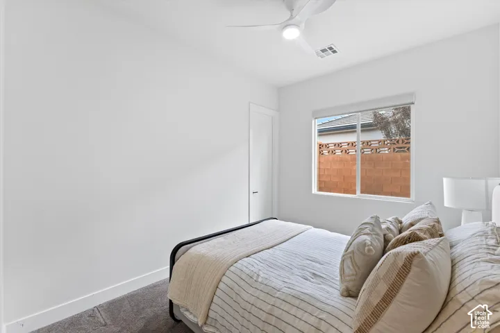 Neutral-toned wall-to-wall carpeting, white interior walls, white ceiling fan with integrated lighting, and single-pane window with exterior brick privacy wall