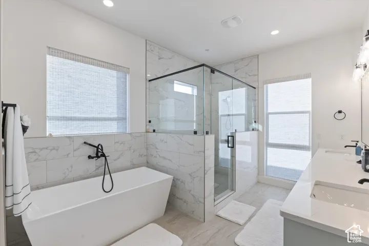 Freestanding soaking tub with wall-mounted matte black faucet, glass-enclosed walk-in shower with stone-finish tile surround, dual-sink vanity with quartz countertop, and recessed lighting