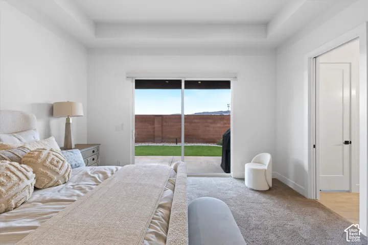 Bedroom featuring a tray ceiling, plush carpeting, and sliding glass doors opening to a concrete patio and fenced backyard