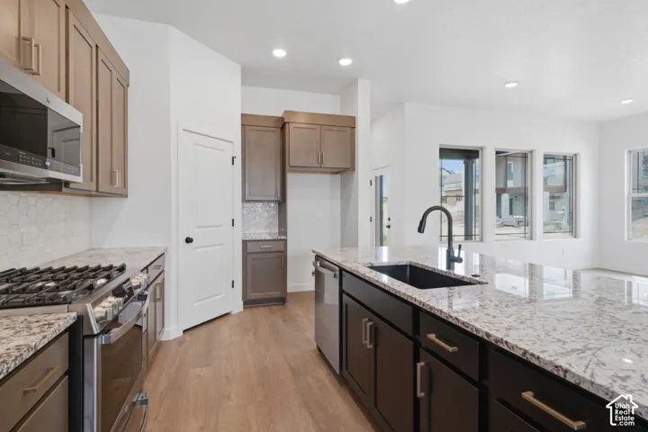 Kitchen featuring light wood-finish cabinetry, stone countertops, hexagonal tile backsplash, a center island with an undermount sink and matte black faucet, and stainless steel appliances