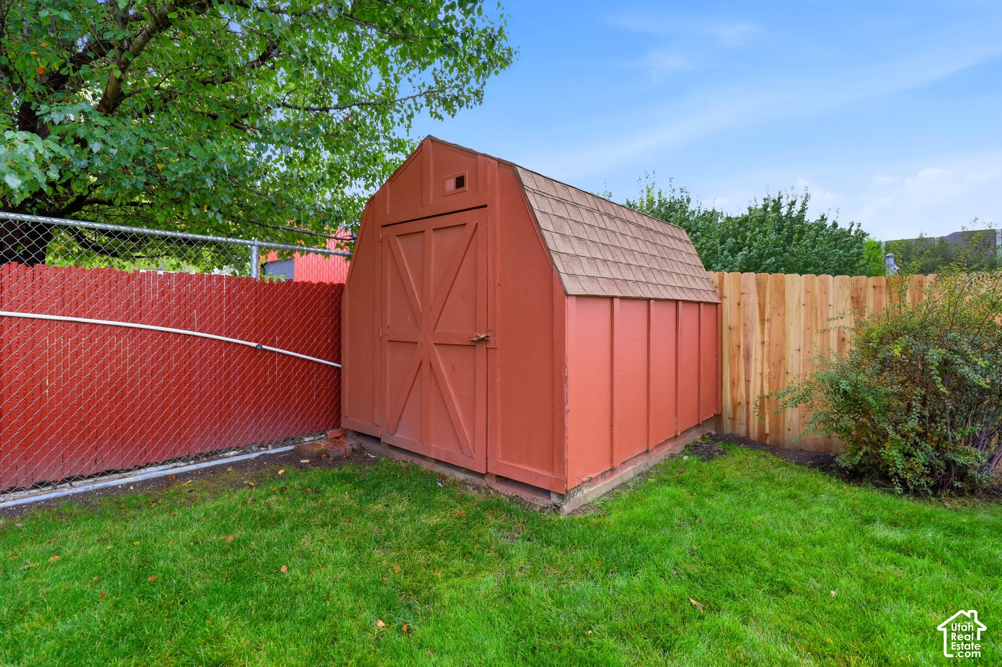 Storage structure with a gambrel roof, shingle roofing, and double-door entry, set within a fenced yard with grass lawn