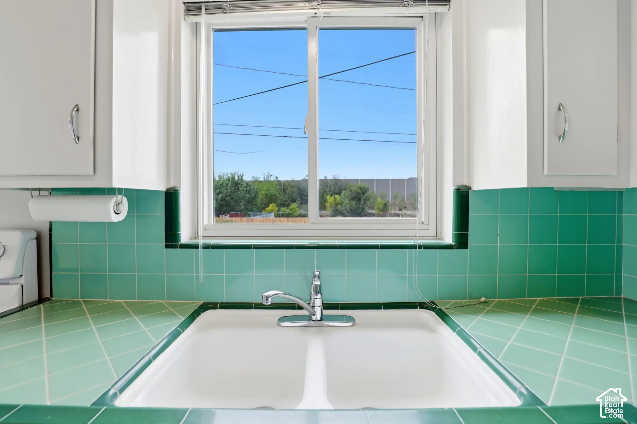 Double-basin sink with a chrome finish faucet, set within green tiled countertops and matching backsplash, framed by white cabinetry and a sliding window