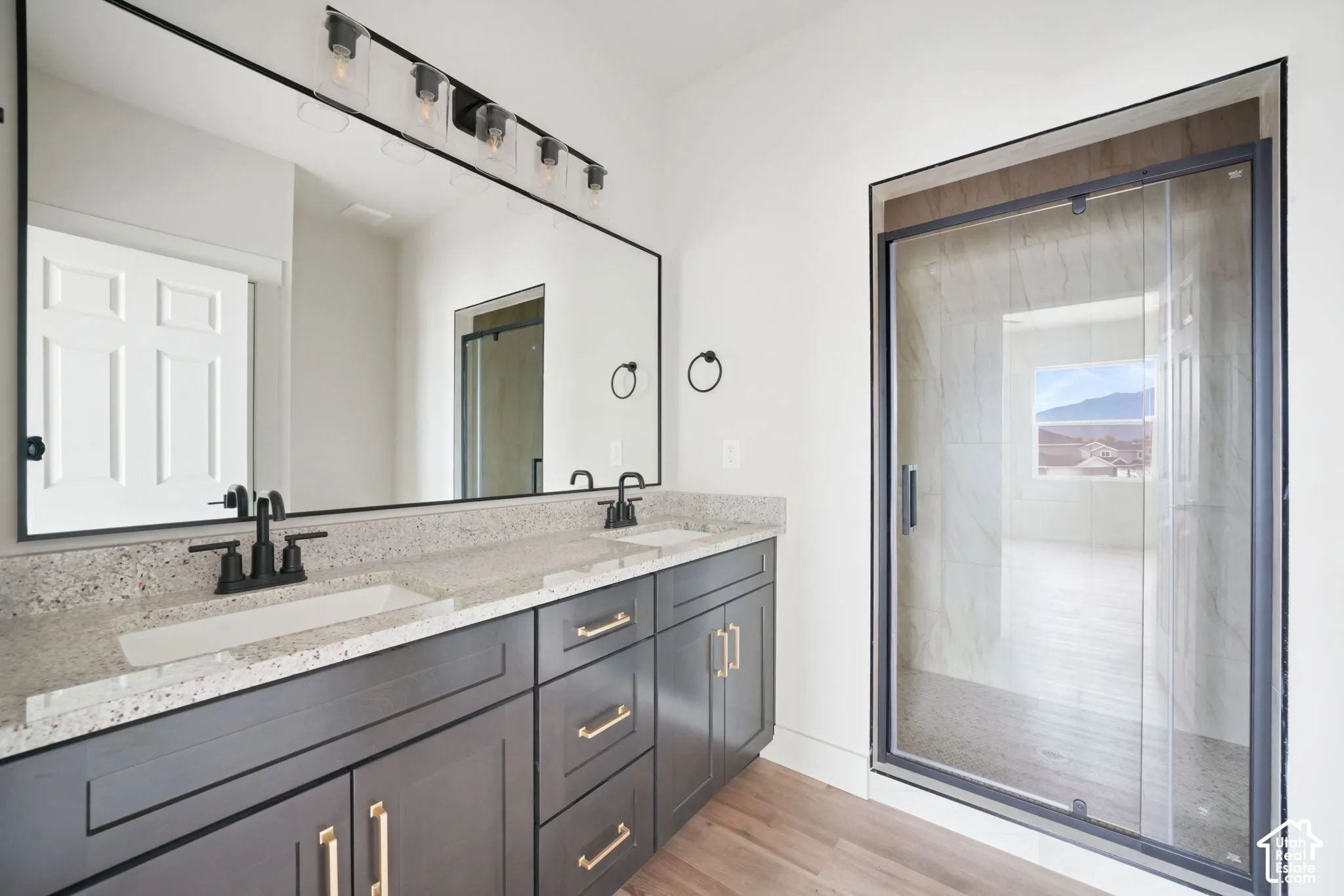 Dual-sink vanity with speckled stone countertop, charcoal cabinetry, matte black hardware, full-width mirror, and walk-in shower with glass enclosure