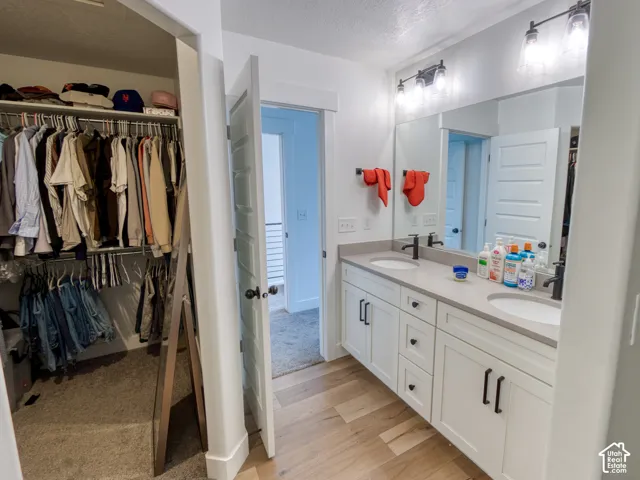Bathroom featuring a dual-sink vanity with matte black hardware, stone-composite countertop, full-width mirror, and wood-finish flooring, complemented by a walk-in closet with built-in shelving