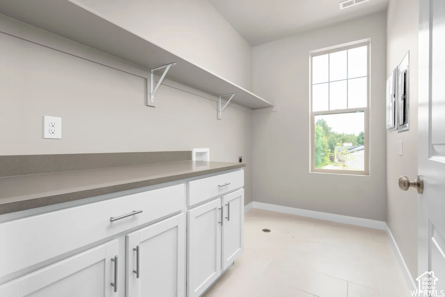 Utility space featuring white cabinetry with brushed-nickel hardware, neutral-toned solid surface countertops, a tall window, and tile flooring