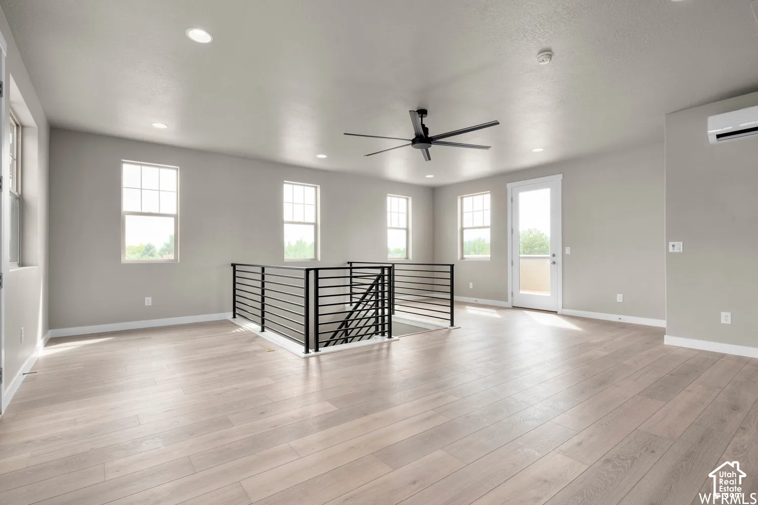 Open-concept space featuring wood-finish flooring, matte black metal railing, recessed lighting, a modern ceiling fan, and a wall-mounted climate control unit