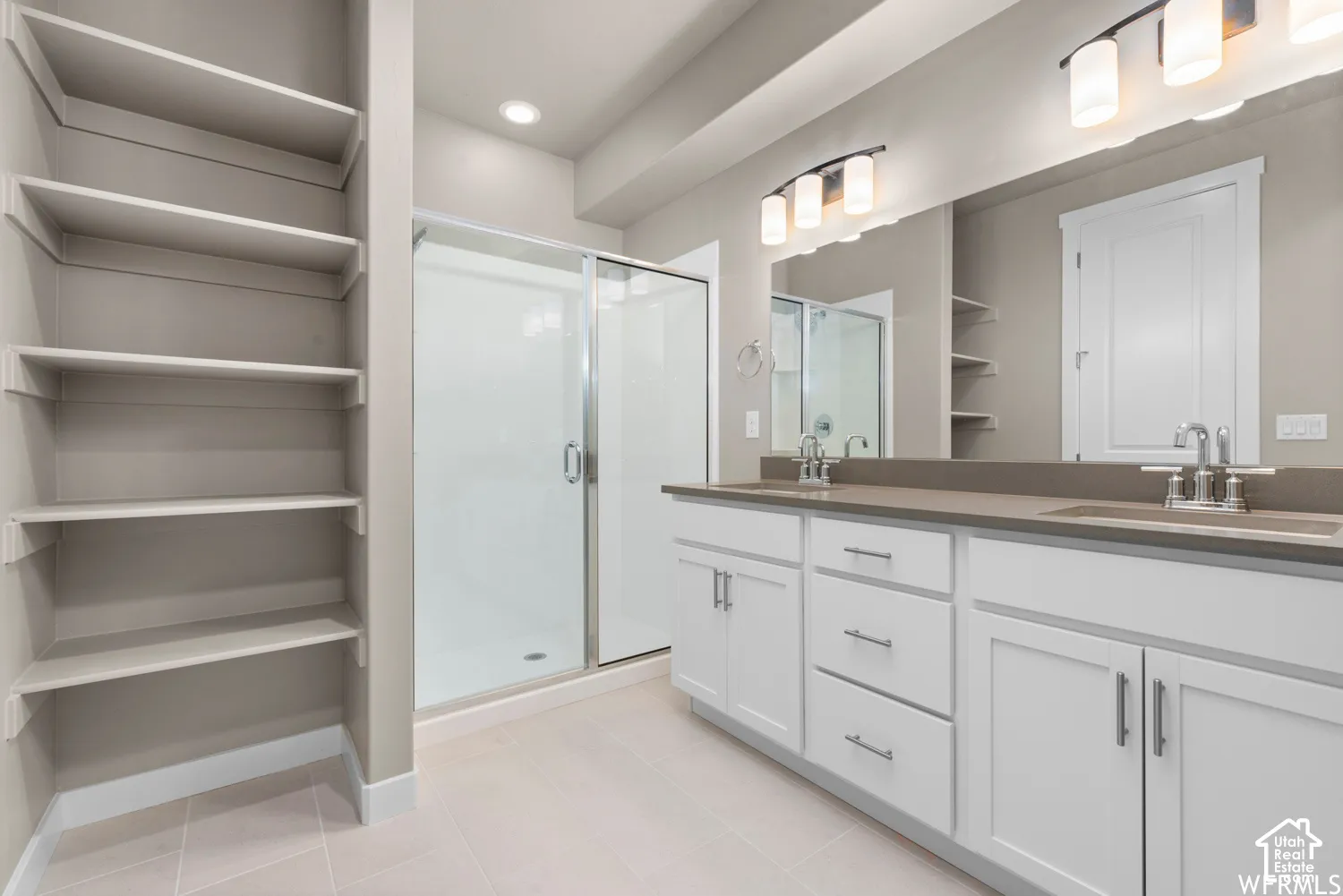 Dual-sink vanity with white cabinetry and solid-surface countertop, walk-in shower with glass enclosure, and built-in shelving unit