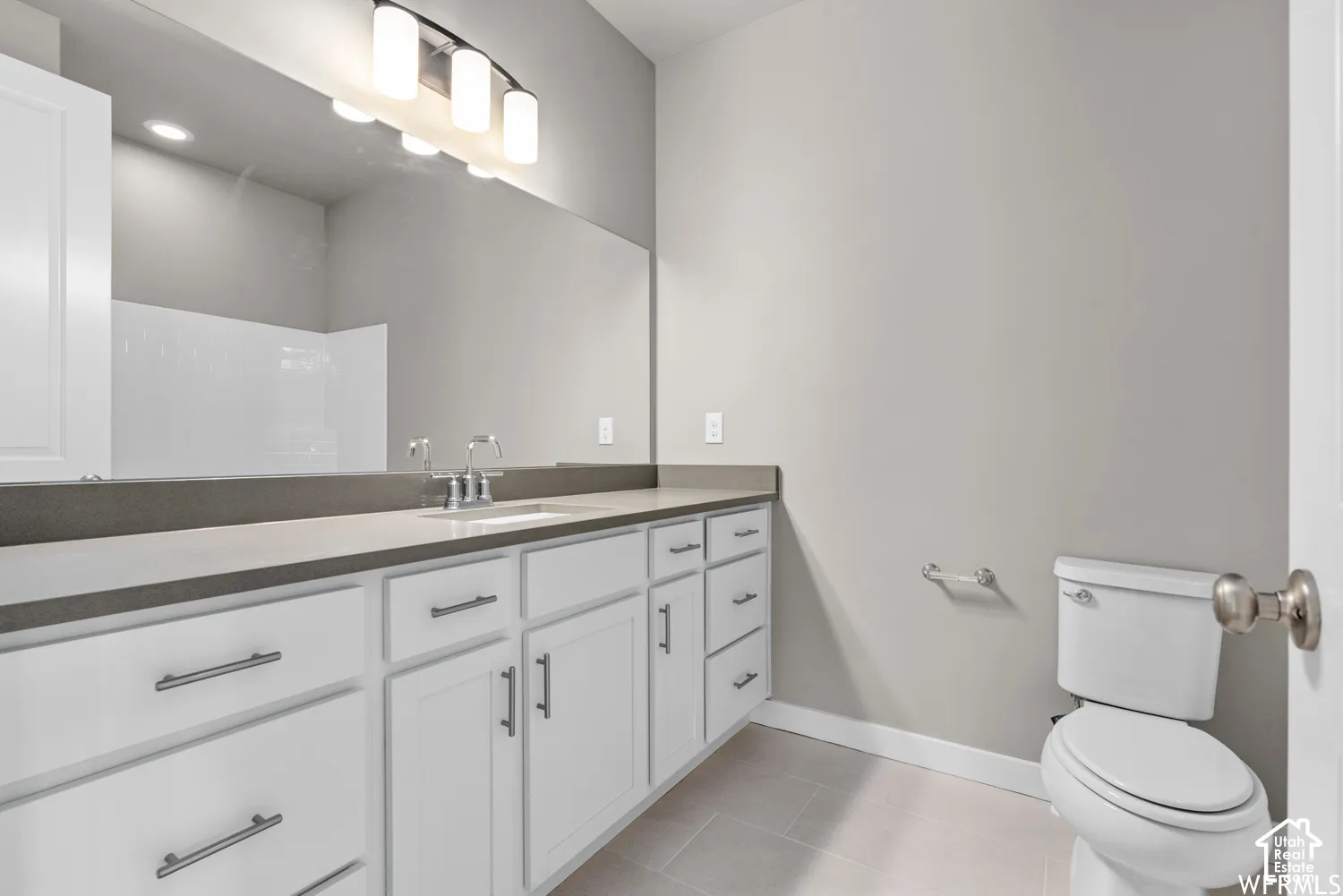 Bathroom featuring a dual-drawer vanity with light-toned countertop, large-format mirror, multi-bulb wall-mounted light fixture, and tile flooring