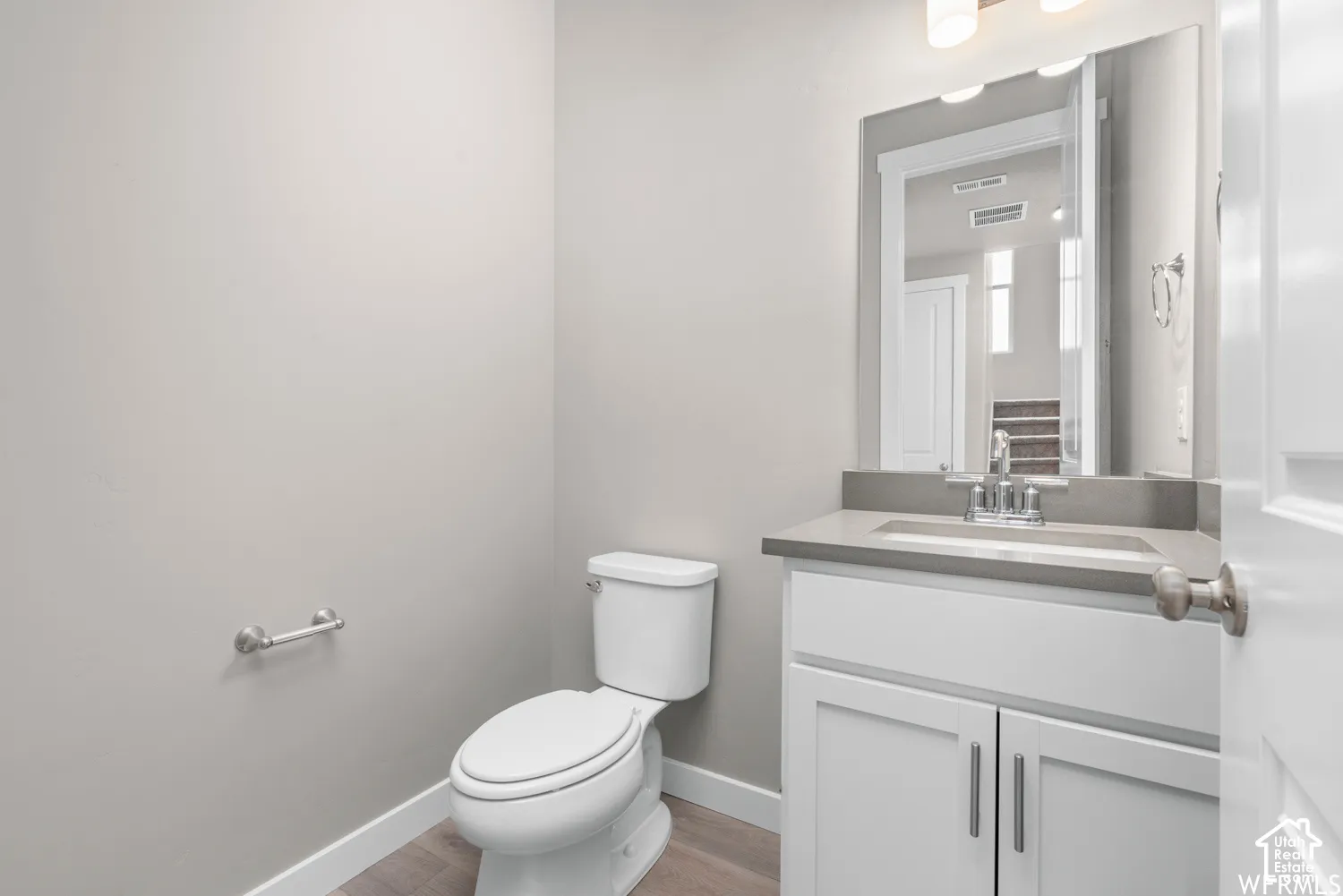 White vanity cabinet with a stone-composite countertop, undermount sink, chrome faucet, wall-mounted mirror, and wood-finish flooring
